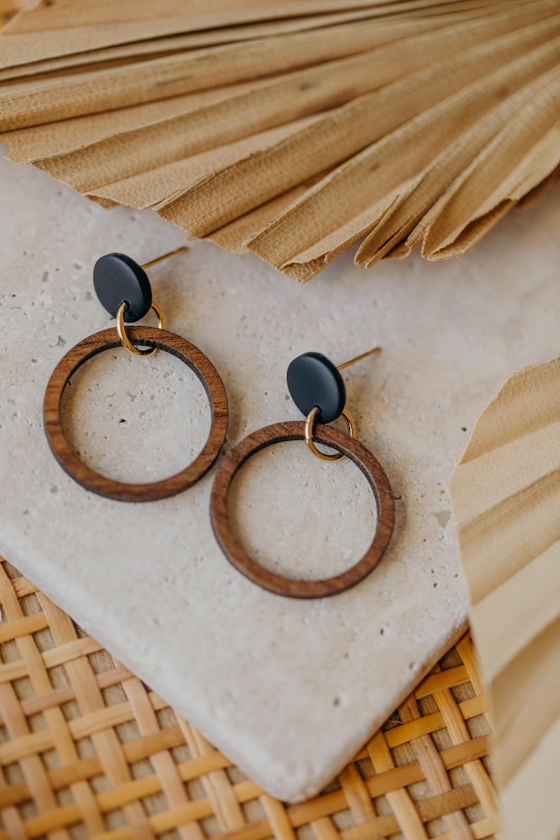 May include: A pair of wooden hoop earrings with a black disc accent and gold-colored hardware. The earrings are displayed on a light-colored stone surface, with dried palm leaves in the background. The earrings have a minimalist, natural aesthetic.