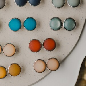 Op de afbeelding: Een assortiment van kleurrijke ronde oorbellen die op een wit oppervlak worden gepresenteerd. De oorbellen zijn gemaakt van verschillende materialen, waaronder metaal, glas en plastic. De oorbellen zijn in paren gerangschikt, waarbij elk paar een andere kleur heeft.