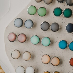 Op de afbeelding: Een assortiment ronde, kleurrijke en metalen oorbelstuds, gepresenteerd op een lichtgrijs oppervlak. De oorbelstuds zijn in paren gerangschikt en hebben verschillende kleuren, waaronder roze, blauw, groen en bruin.