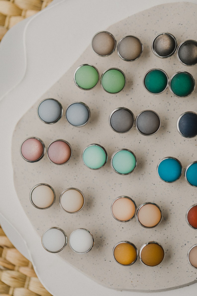 Op de afbeelding: Een assortiment ronde, kleurrijke en metalen oorbelstuds, gepresenteerd op een lichtgrijs oppervlak. De oorbelstuds zijn in paren gerangschikt en hebben verschillende kleuren, waaronder roze, blauw, groen en bruin.