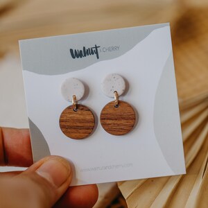 May include: A pair of earrings with a white speckled clay top and a round wooden bottom. The earrings are displayed on a white card with the text "Walnut & Cherry" and a website address.
