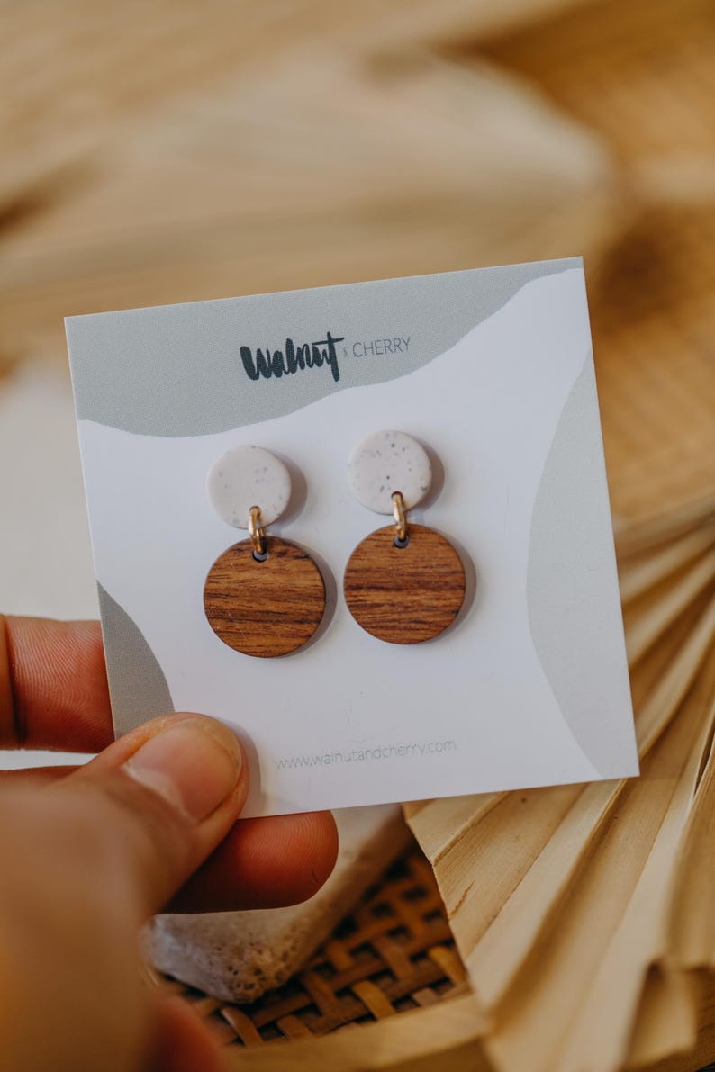 May include: A pair of earrings with a white speckled clay top and a round wooden bottom. The earrings are displayed on a white card with the text "Walnut & Cherry" and a website address.