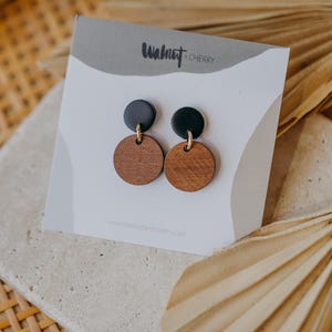 May include: A pair of earrings with black and brown round wooden discs. The earrings are on a white card with the text "Walnut + Cherry" printed on it.