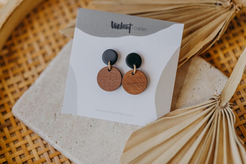 May include: A pair of earrings with black and brown round wooden discs. The earrings are on a white card with the text "Walnut + Cherry" printed on it.