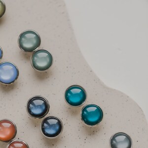 Op de afbeelding: Assortiment van ronde, gekleurde glazen cabochons met zilveren instellingen. De cabochons zijn gerangschikt in rijen op een wit oppervlak.