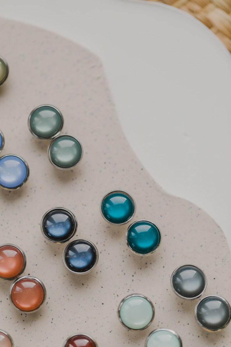 Op de afbeelding: Assortiment van ronde, gekleurde glazen cabochons met zilveren instellingen. De cabochons zijn gerangschikt in rijen op een wit oppervlak.
