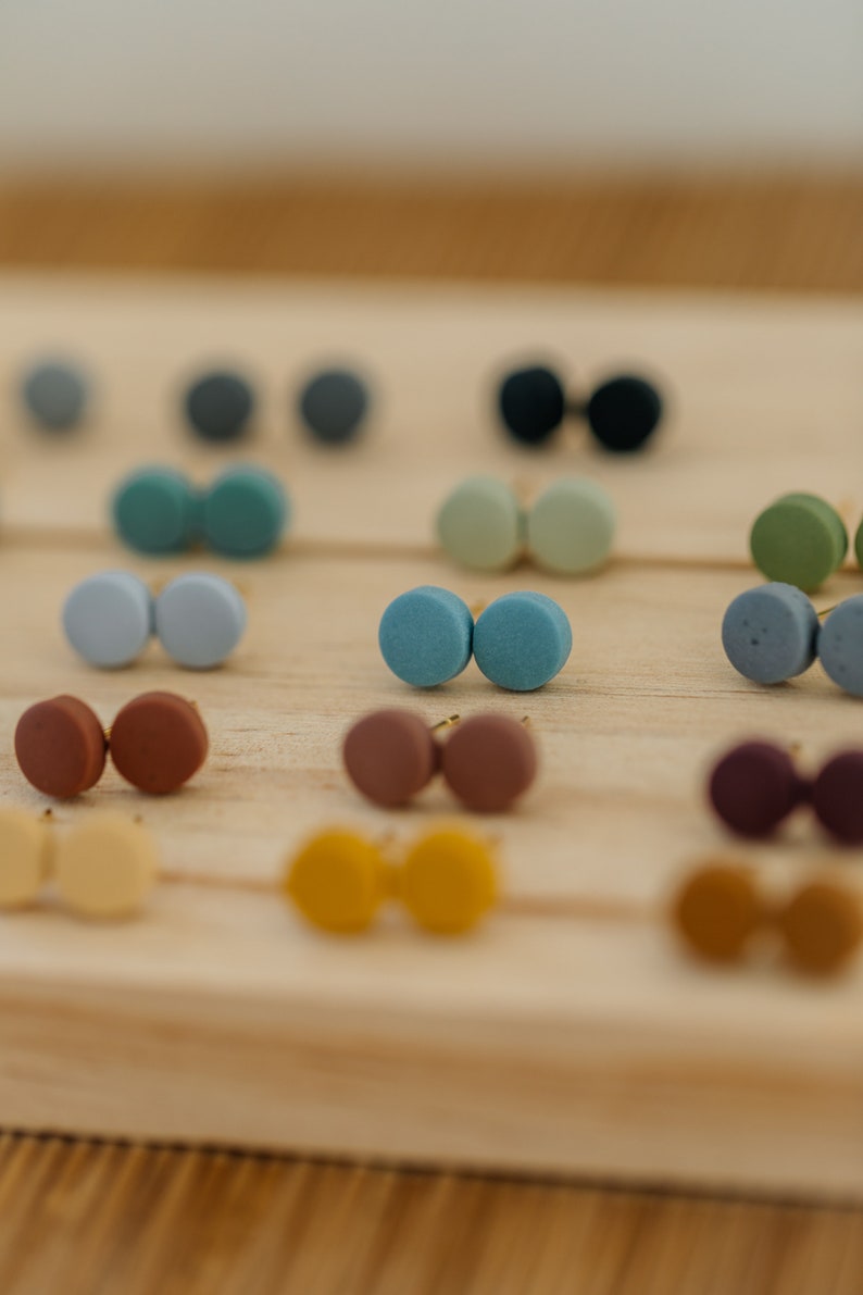May include: A variety of small, round, colorful earrings displayed on a wooden surface. The earrings are made of clay and come in pairs.