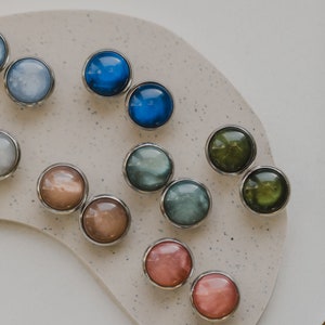 May include: Assortment of colorful round buttons with silver settings. The buttons are arranged in a semi-circle on a light brown surface.