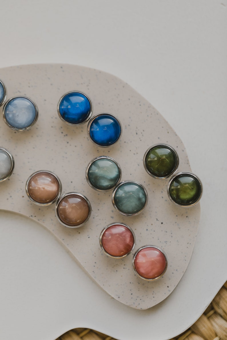 May include: Assortment of colorful round buttons with silver settings. The buttons are arranged in a semi-circle on a light brown surface.
