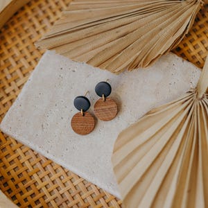 May include: A pair of earrings with black and brown round wooden discs. The earrings are displayed on a white tile surface with a woven basket and palm leaves in the background.