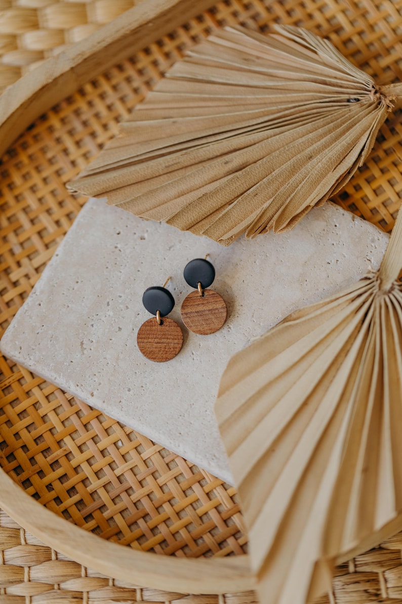 May include: A pair of earrings with black and brown round wooden discs. The earrings are displayed on a white tile surface with a woven basket and palm leaves in the background.