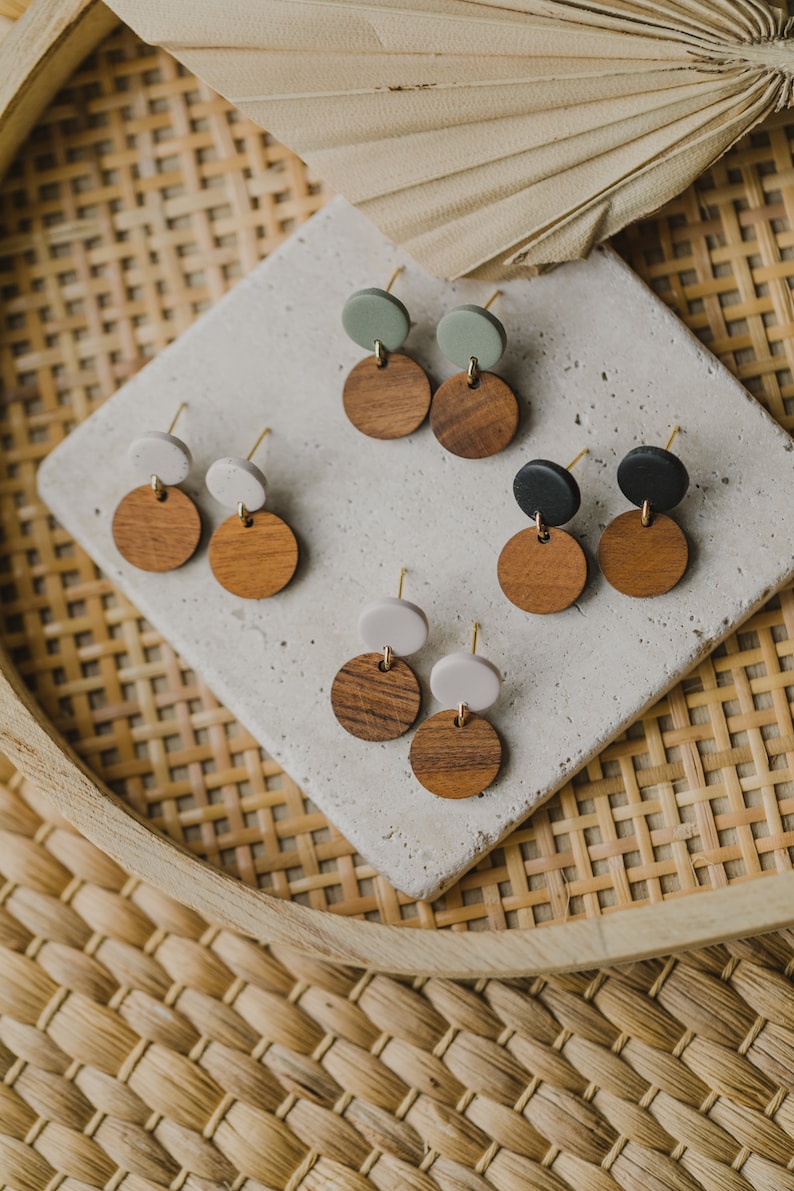May include: Six pairs of earrings with two-toned round wooden and colored clay discs. The earrings are displayed on a white surface with a woven background.