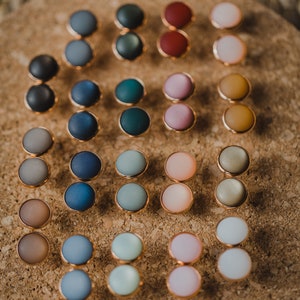 Pu&ograve; includere: Un set di 20 paia di orecchini rotondi e colorati. Gli orecchini sono realizzati in un materiale liscio e opaco e sono disposti in una griglia su una superficie di sughero.