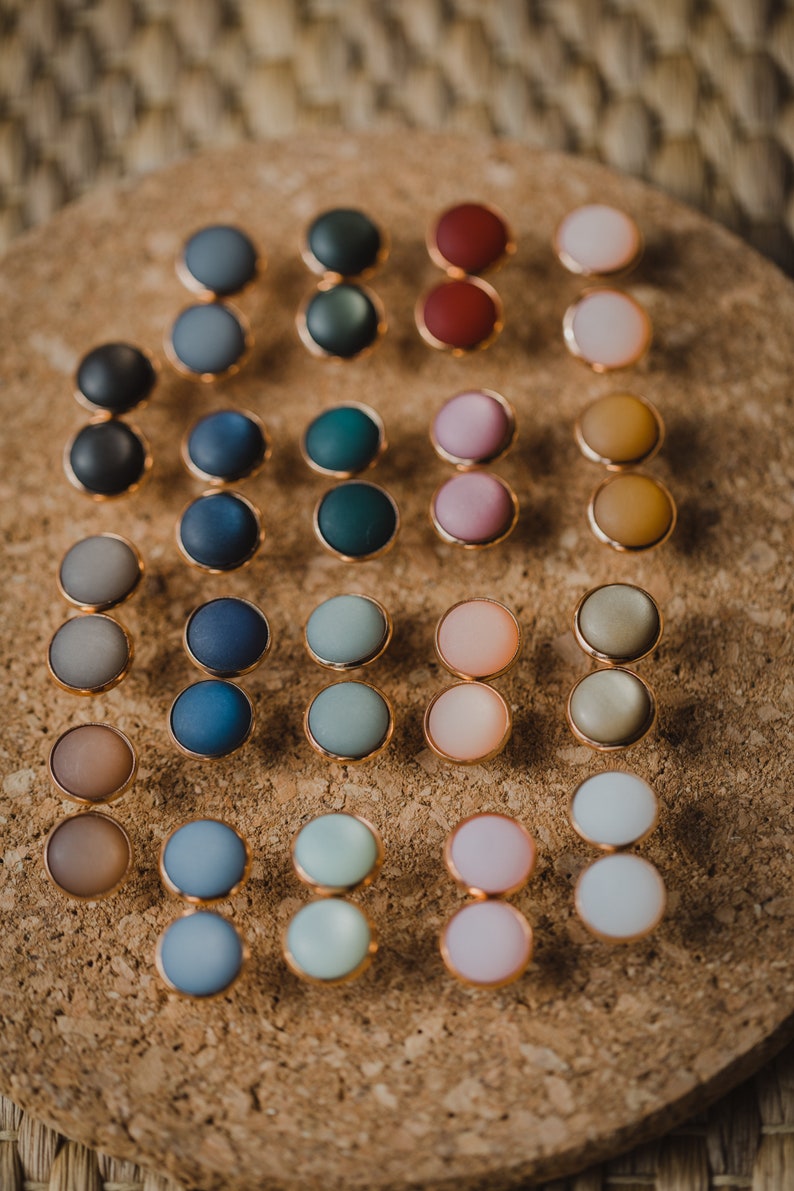 Pu&ograve; includere: Un set di 20 paia di orecchini rotondi e colorati. Gli orecchini sono realizzati in un materiale liscio e opaco e sono disposti in una griglia su una superficie di sughero.