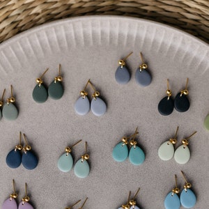 May include: A collection of 18 pairs of handmade ceramic earrings in various shades of blue, green, and gray. Each earring features a teardrop shape with a gold accent.