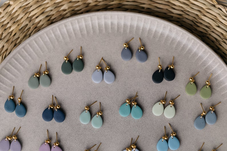 May include: A collection of 18 pairs of handmade ceramic earrings in various shades of blue, green, and gray. Each earring features a teardrop shape with a gold accent.