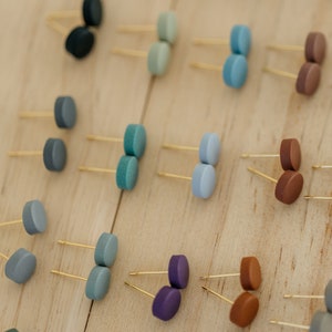 May include: A collection of small, round, colorful earrings displayed on a wooden surface. The earrings are made of polymer clay and come in a variety of colors, including blue, green, purple, brown, and gray.