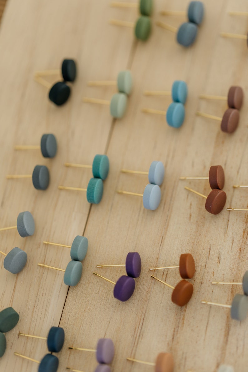 May include: A collection of small, round, colorful earrings displayed on a wooden surface. The earrings are made of polymer clay and come in a variety of colors, including blue, green, purple, brown, and gray.