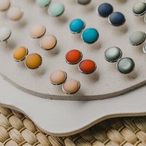 Op de afbeelding: Een display met ronde oorbellen in verschillende kleuren op een wit platform. De oorbellen zijn in paren gerangschikt en verkrijgbaar in verschillende kleuren, waaronder blauw, groen, rood, oranje en roze.