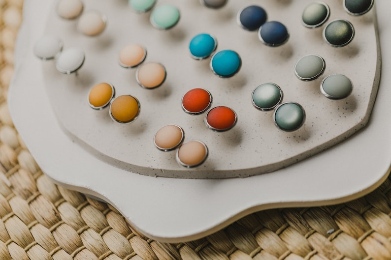 Op de afbeelding: Een display met ronde oorbellen in verschillende kleuren op een wit platform. De oorbellen zijn in paren gerangschikt en verkrijgbaar in verschillende kleuren, waaronder blauw, groen, rood, oranje en roze.