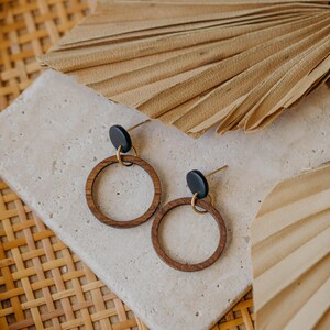 May include: A pair of wooden hoop earrings with black studs and gold-colored posts. The earrings are displayed on a light-colored stone surface, with a woven basket and dried palm leaves in the background.