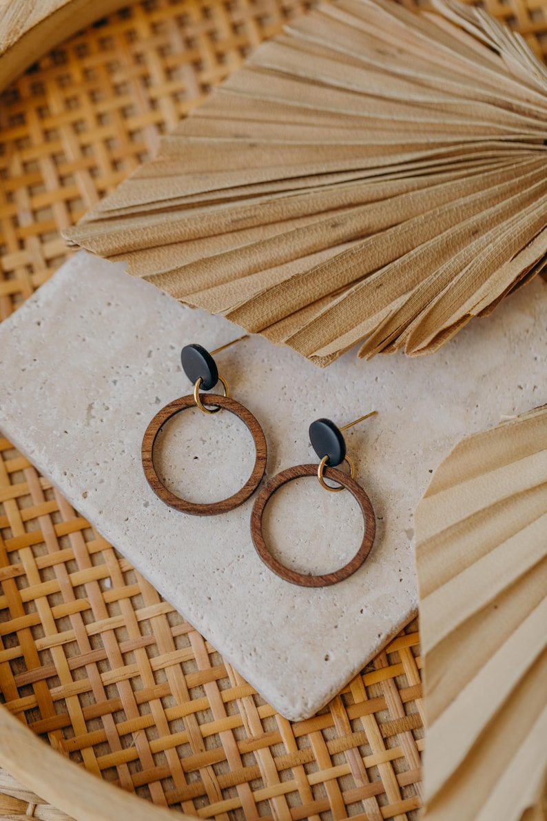 May include: A pair of wooden hoop earrings with black studs and gold-colored posts. The earrings are displayed on a light-colored stone surface, with a woven basket and dried palm leaves in the background.