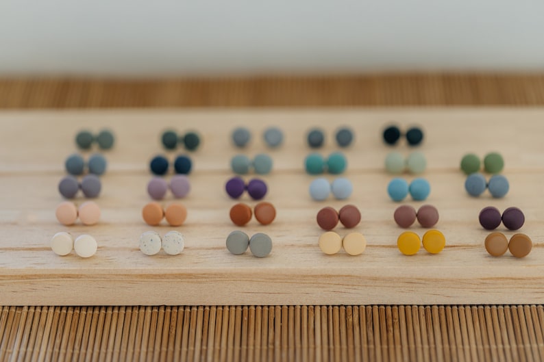 May include: A collection of 20 pairs of small, round, colorful clay earrings displayed on a wooden board. The earrings are arranged in rows of two, with each pair featuring a different color or pattern.