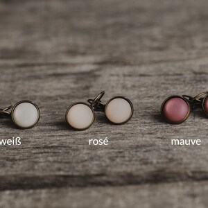 May include: Three pairs of earrings with different coloured cabochons. The earrings are white, rose, and mauve. The earrings are on a wooden surface.