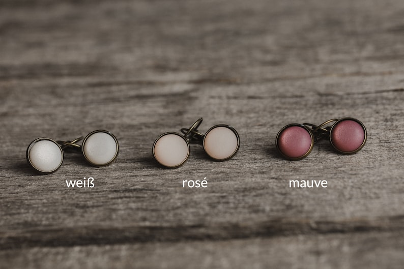 May include: Three pairs of earrings with different coloured cabochons. The earrings are white, rose, and mauve. The earrings are on a wooden surface.
