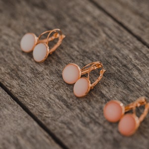 May include: Three pairs of gold hoop earrings with round white and pink stones. The earrings are on a wooden surface.