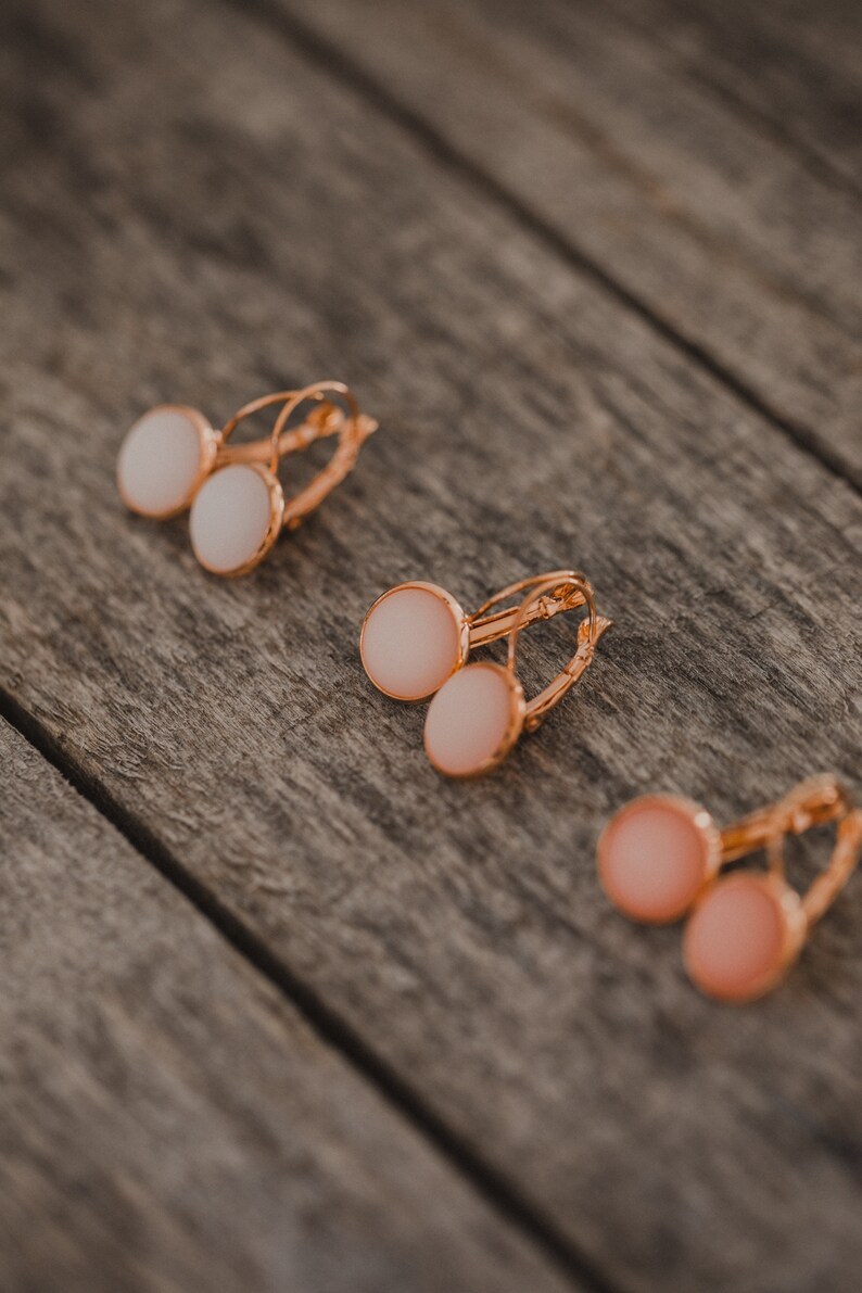 May include: Three pairs of gold hoop earrings with round white and pink stones. The earrings are on a wooden surface.