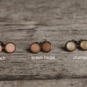 May include: Close-up of six small round earrings in three different colours: peach, brown-taupe, and champagne. The earrings are displayed on a wooden surface.