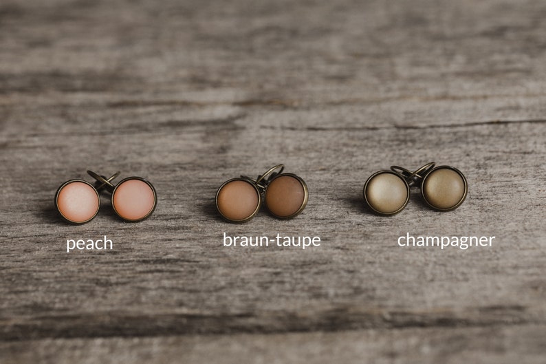 May include: Close-up of six small round earrings in three different colours: peach, brown-taupe, and champagne. The earrings are displayed on a wooden surface.