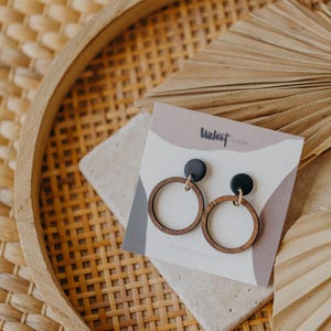 May include: Wooden hoop earrings with black tops and gold-colored hooks, displayed on a neutral-toned card. The card has the text "Walnut Cherie" in a handwritten font. The earrings are set on a stone-like surface within a woven tray.