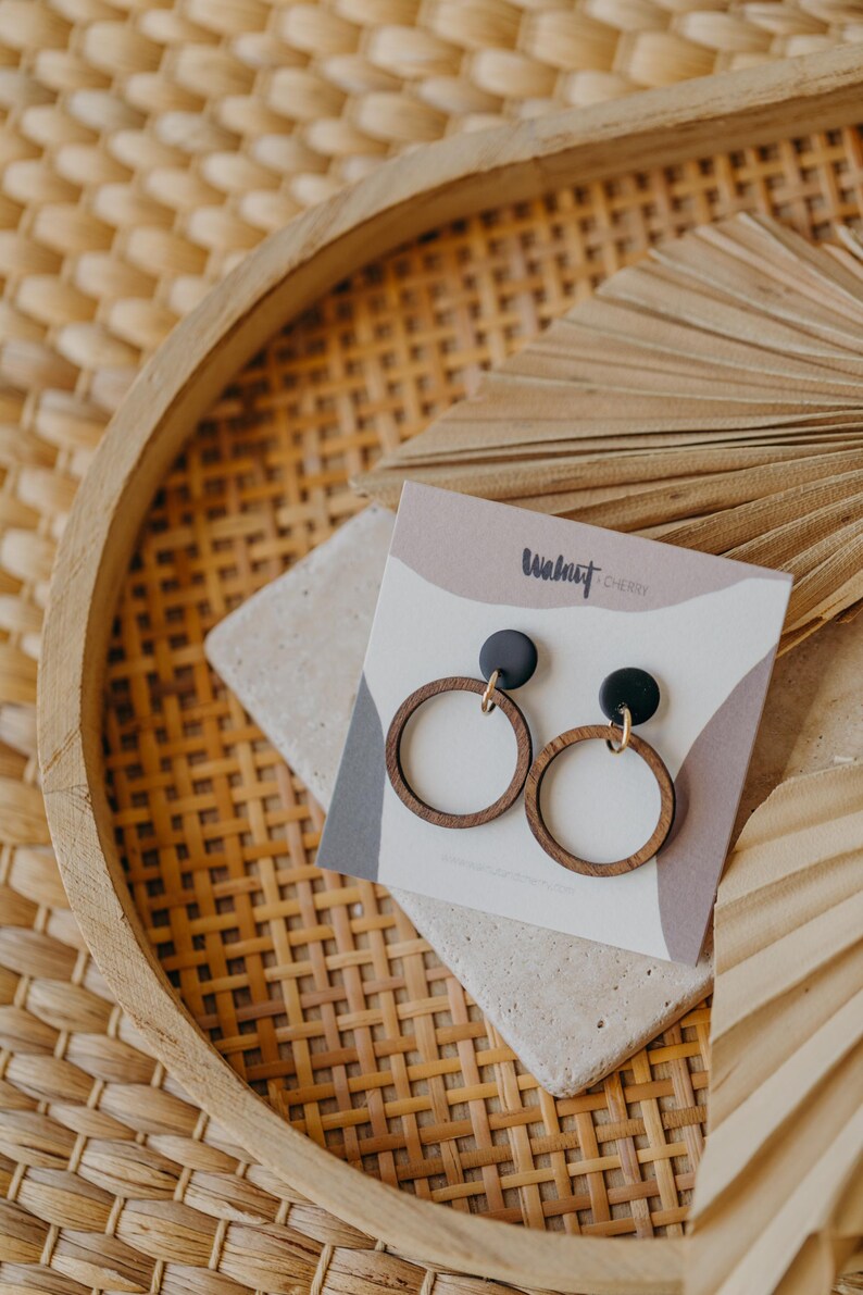 May include: Wooden hoop earrings with black tops and gold-colored hooks, displayed on a neutral-toned card. The card has the text "Walnut Cherie" in a handwritten font. The earrings are set on a stone-like surface within a woven tray.