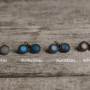 May include: Six pairs of small, round earrings with different coloured glass inlays. The colours are grey-blue, dark blue, jeans blue, and chocolate brown.