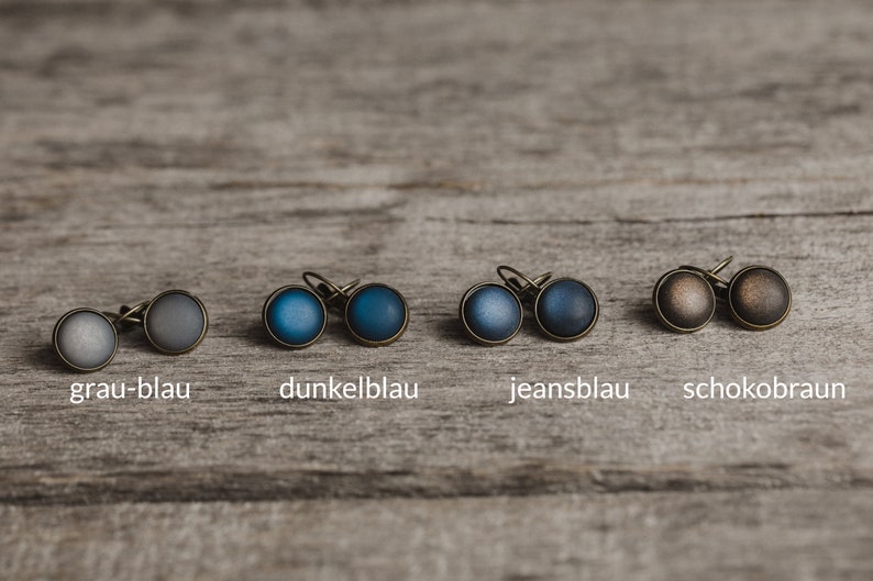 May include: Six pairs of small, round earrings with different coloured glass inlays. The colours are grey-blue, dark blue, jeans blue, and chocolate brown.