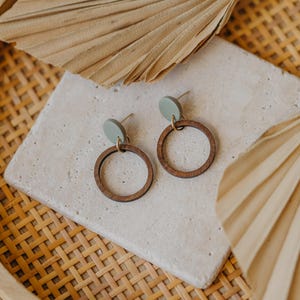 May include: A pair of wooden hoop earrings with a light green circle accent. The earrings are displayed on a white tile surface with a woven background.