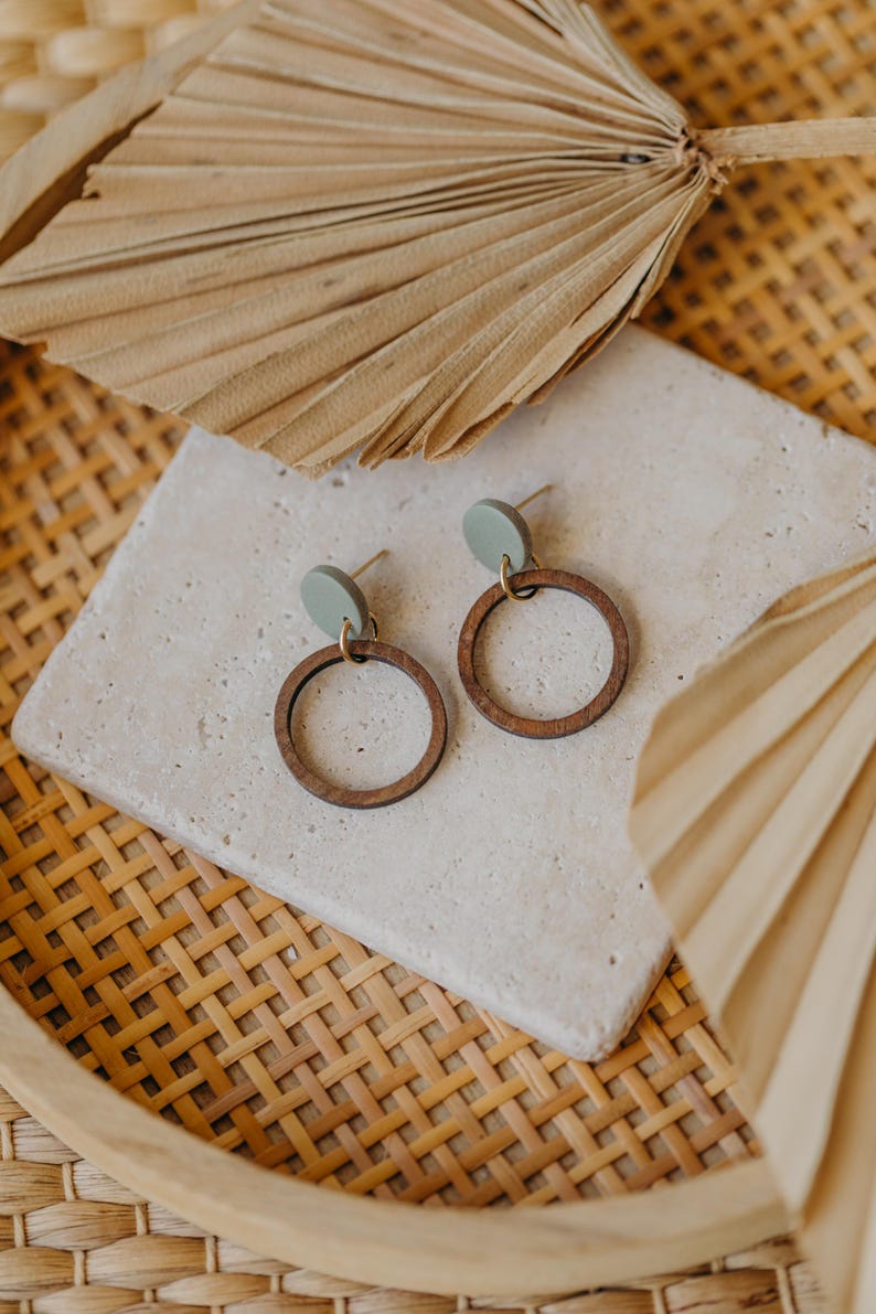 May include: A pair of wooden hoop earrings with a light green circle accent. The earrings are displayed on a white tile surface with a woven background.