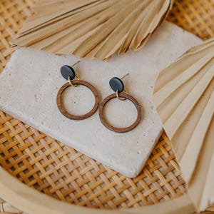 May include: A pair of wooden hoop earrings with black studs and gold-colored posts. The earrings are displayed on a light-colored stone, with dried palm leaves and a woven tray in the background. The earrings have a minimalist, bohemian style.
