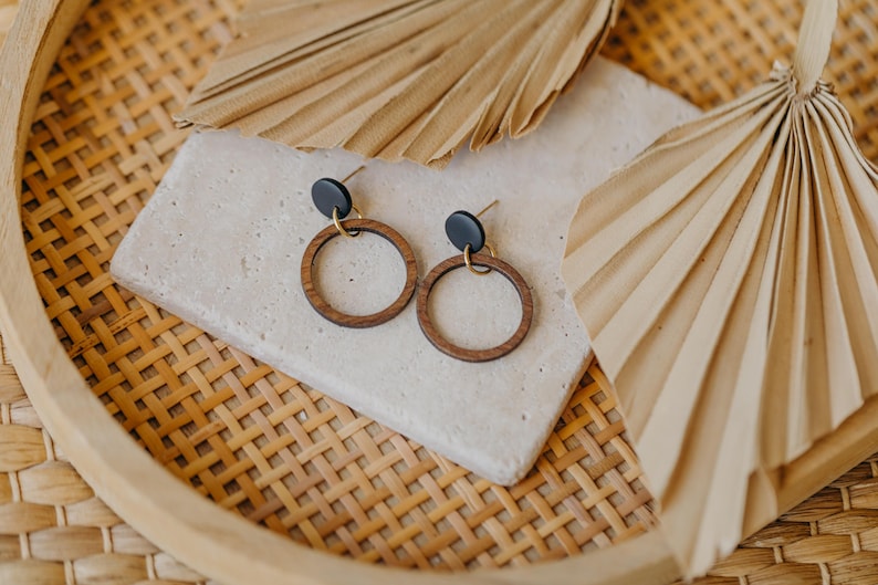 May include: A pair of wooden hoop earrings with black studs and gold-colored posts. The earrings are displayed on a light-colored stone, with dried palm leaves and a woven tray in the background. The earrings have a minimalist, bohemian style.