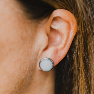 Op de afbeelding: Een close-up van iemands oor met een witte ronde oorbel. De oorbel is gemaakt van glad, wit materiaal en heeft een zilveren metalen achterkant.