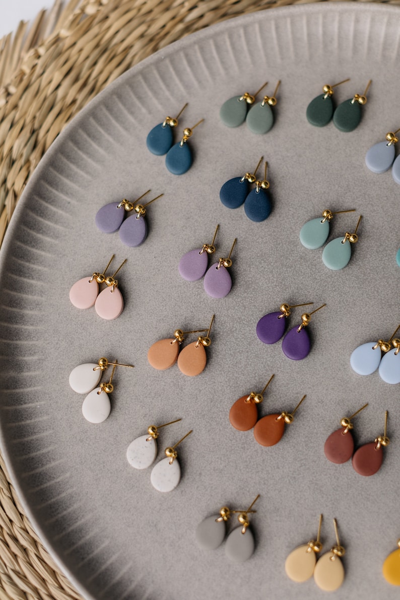 May include: A collection of 18 pairs of teardrop-shaped earrings in various colors, including blue, green, purple, pink, brown, gray, and white. The earrings are made of clay and have gold-colored posts.