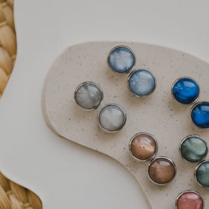 May include: Assortment of round, colorful, iridescent buttons with silver settings. The buttons are arranged on a speckled, light brown surface.