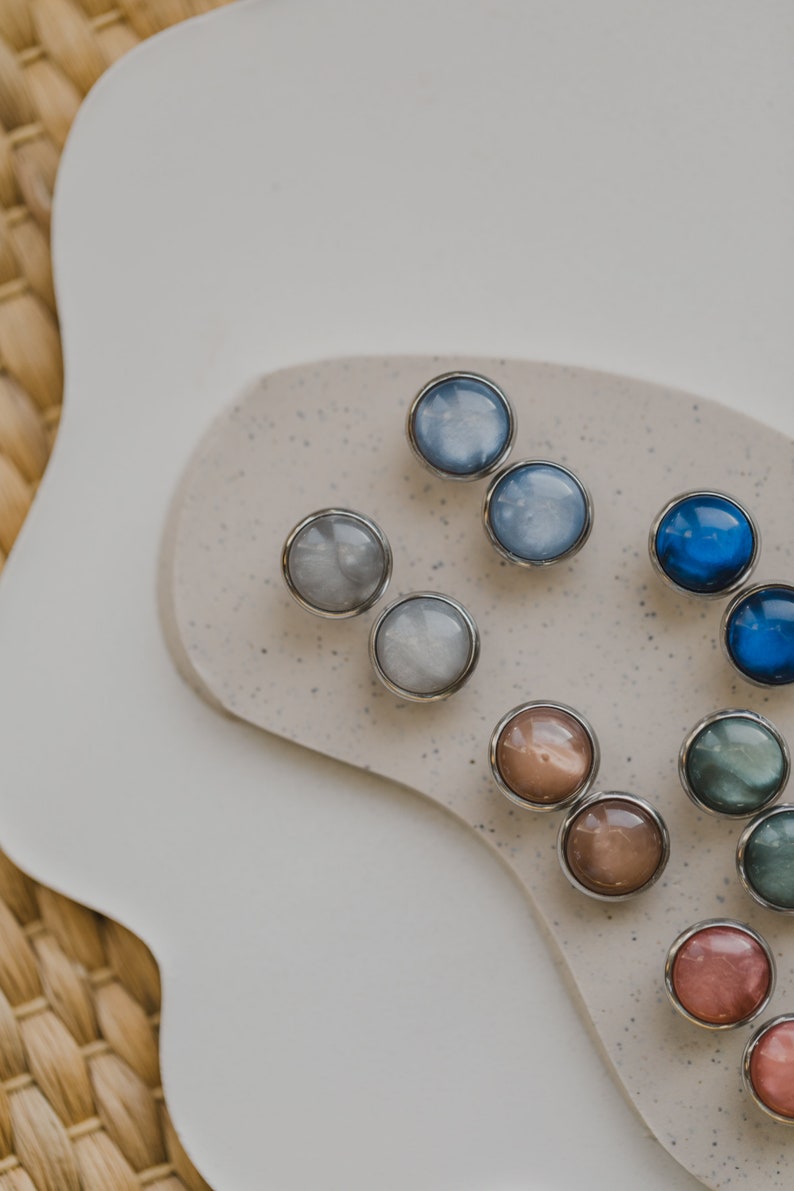 May include: Assortment of round, colorful, iridescent buttons with silver settings. The buttons are arranged on a speckled, light brown surface.