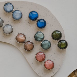 May include: Assortment of round, iridescent buttons in various colors, including blue, green, pink, and brown. The buttons are arranged in pairs on a light beige surface.