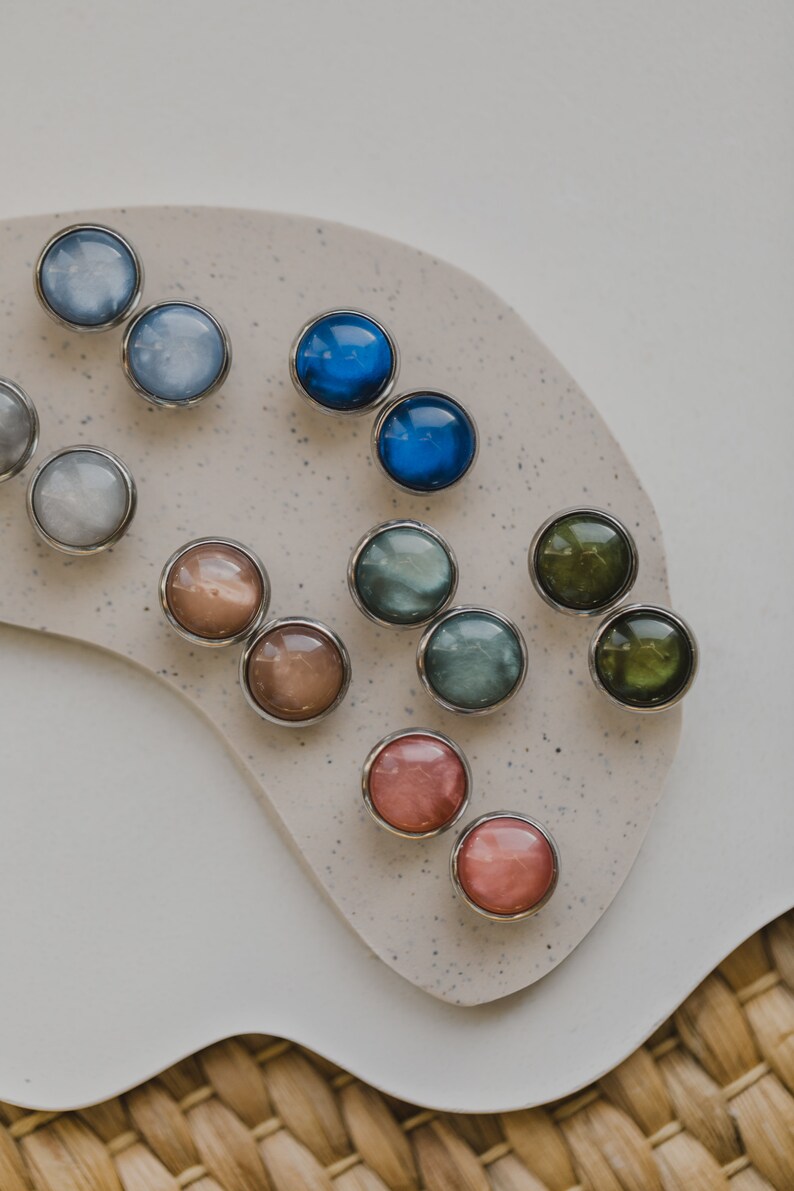 May include: Assortment of round, iridescent buttons in various colors, including blue, green, pink, and brown. The buttons are arranged in pairs on a light beige surface.