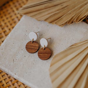 May include: A pair of wooden and white circle earrings. The earrings are small and have a simple design. The wooden circles are a dark brown color and the white circles are a light cream color.
