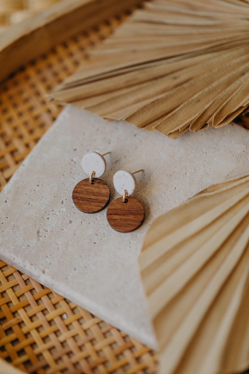 May include: A pair of wooden and white circle earrings. The earrings are small and have a simple design. The wooden circles are a dark brown color and the white circles are a light cream color.
