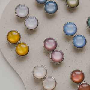 Op de afbeelding: Assortiment van ronde glazen cabochons in verschillende kleuren met zilveren instellingen. De cabochons zijn in paren gerangschikt op een lichtgrijs oppervlak.